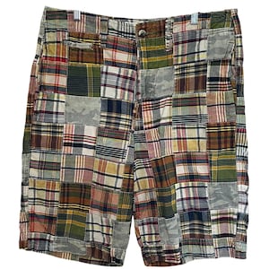 May include: Patchwork shorts featuring a variety of plaid and camouflage fabric squares in shades of olive green, navy blue, red, yellow, and white. The shorts include a button closure at the waist, belt loops, and side pockets with a visible seam construction.