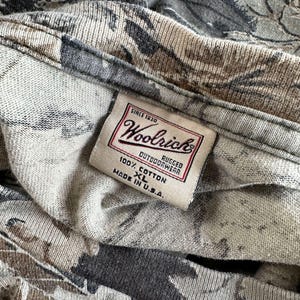 Puede incluir: Primer plano de una camisa con estampado de camuflaje con una etiqueta Woolrich. La etiqueta dice "SINCE 1830 Woolrich RUGGED OUTDOORWEAR 100% COTTON XL MADE IN U.S.A." La camisa es de algod&oacute;n y presenta una mezcla de colores grises, marrones y negros.