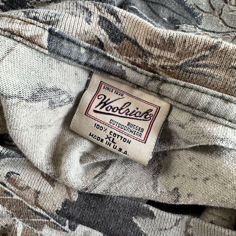 Puede incluir: Primer plano de una camisa con estampado de camuflaje con una etiqueta Woolrich. La etiqueta dice "SINCE 1830 Woolrich RUGGED OUTDOORWEAR 100% COTTON XL MADE IN U.S.A." La camisa es de algod&oacute;n y presenta una mezcla de colores grises, marrones y negros.