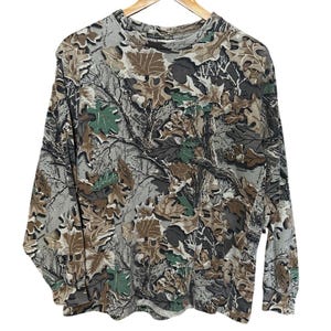 Puede incluir: Camiseta de camuflaje de manga larga con cuello redondo. La camiseta presenta un estampado de hojas y ramas marrones, verdes y grises. La camiseta est&aacute; colgada de una percha de madera.