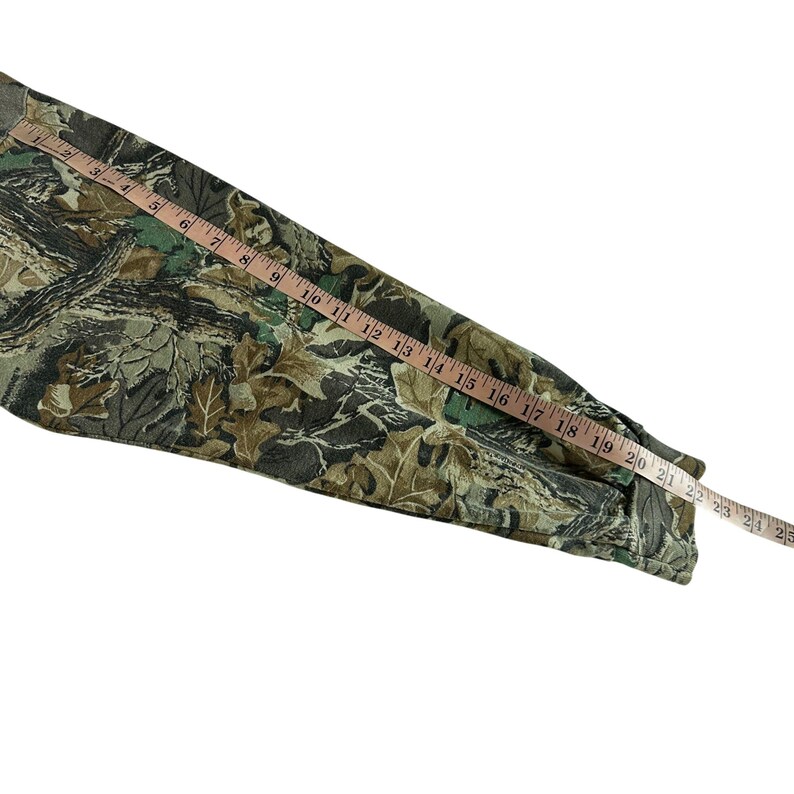 Puede incluir: Una camisa de manga larga con estampado de camuflaje con una cinta m&eacute;trica colocada encima. La camisa presenta un dise&ntilde;o de hojas verdes, marrones y negras. La cinta m&eacute;trica muestra medidas de hasta 63,5 cm.