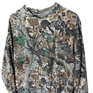 Puede incluir: Camiseta de camuflaje de manga larga con cuello alto. La camiseta presenta un estampado de hojas y ramas marrones, verdes y grises. La tela parece ser un material suave y c&oacute;modo.
