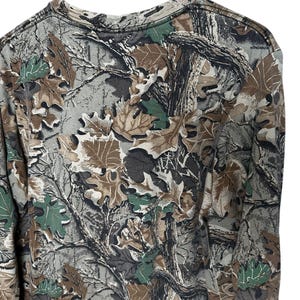 Puede incluir: Una camiseta de manga larga de camuflaje con un estampado de hojas y ramas marrones, verdes y grises. La camiseta tiene cuello redondo y est&aacute; hecha de un material suave. El dise&ntilde;o es un camuflaje de bosque cl&aacute;sico.