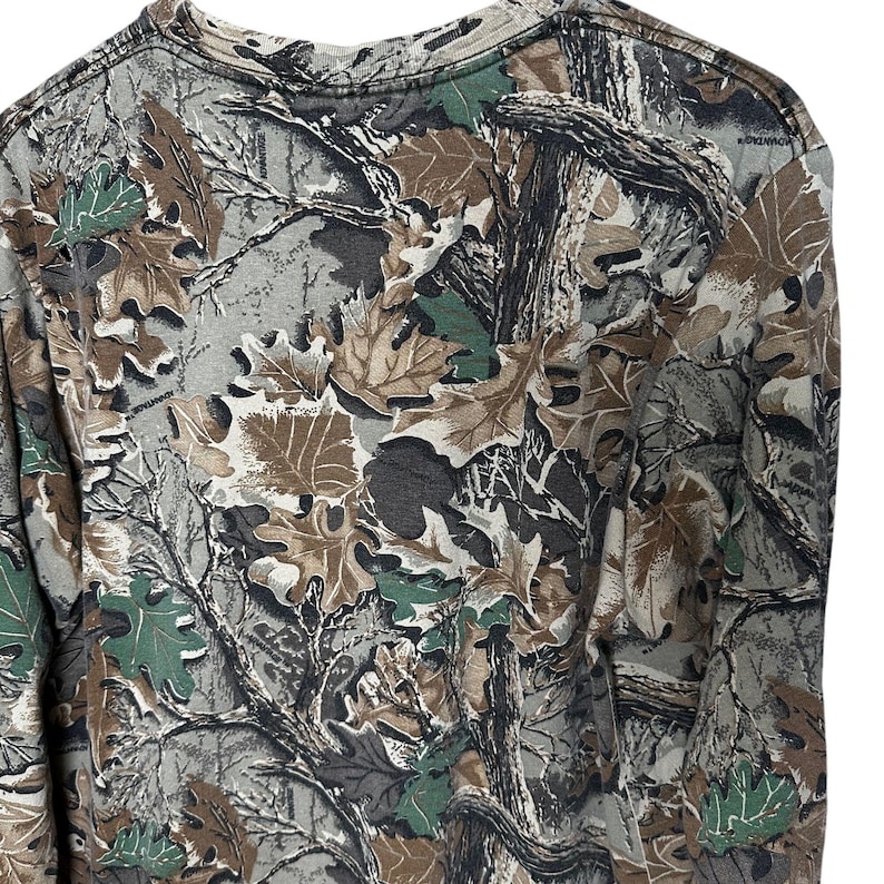Puede incluir: Una camiseta de manga larga de camuflaje con un estampado de hojas y ramas marrones, verdes y grises. La camiseta tiene cuello redondo y est&aacute; hecha de un material suave. El dise&ntilde;o es un camuflaje de bosque cl&aacute;sico.