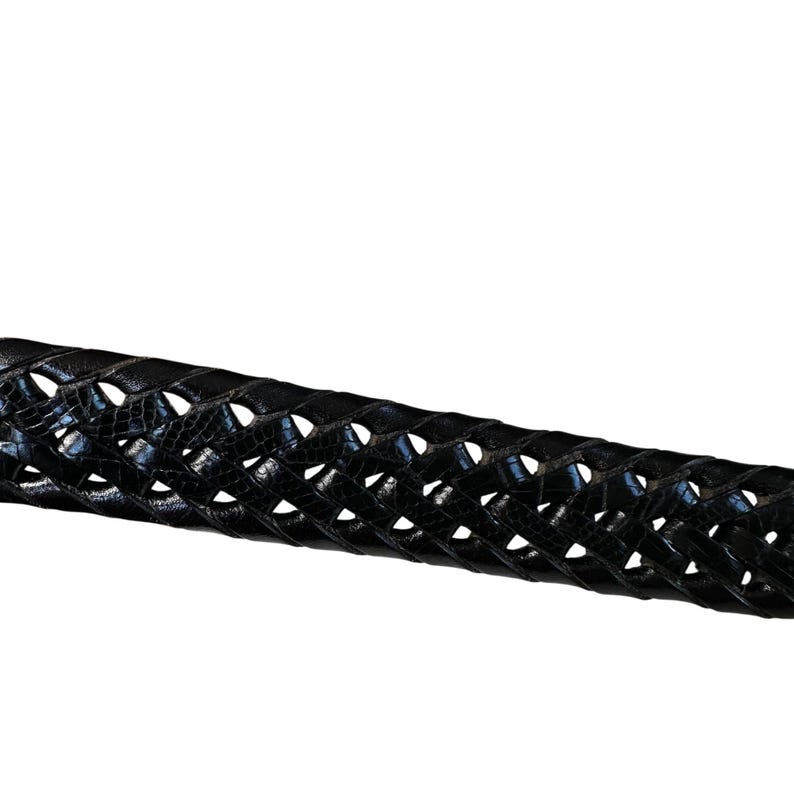 May include: Close-up of a black, braided leather belt. The belt has a textured, crocodile-like pattern and is woven in a repeating pattern. The belt is a dark, glossy black.