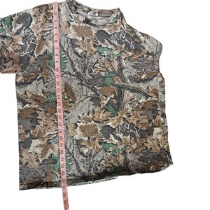 Puede incluir: Una camiseta con estampado de camuflaje con cuello redondo. La camiseta presenta un dise&ntilde;o de hojas y ramas marrones, verdes y grises. Una cinta m&eacute;trica rosa se coloca verticalmente a lo largo de la camiseta, mostrando las medidas en cent&iacute;metros.
