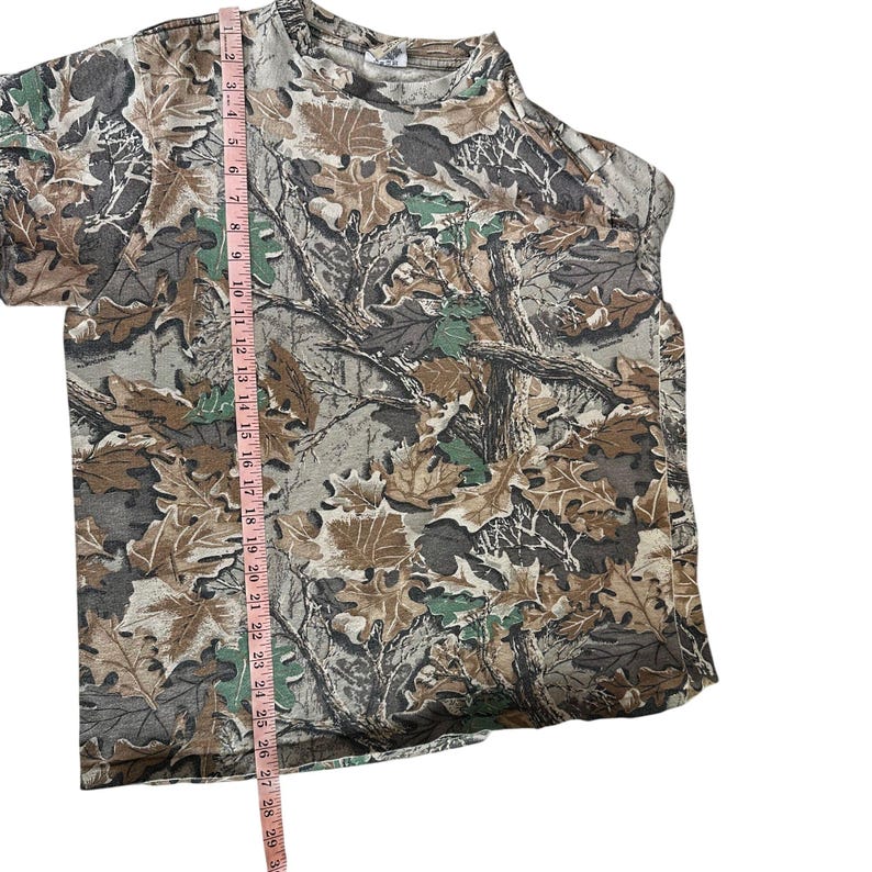 Puede incluir: Una camiseta con estampado de camuflaje con cuello redondo. La camiseta presenta un dise&ntilde;o de hojas y ramas marrones, verdes y grises. Una cinta m&eacute;trica rosa se coloca verticalmente a lo largo de la camiseta, mostrando las medidas en cent&iacute;metros.