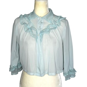 May include: A light blue vintage blouse with a collar and ruffled trim. The blouse has three-quarter sleeves with ruffled cuffs and a button-down front. The fabric appears to be a lightweight, semi-sheer material.