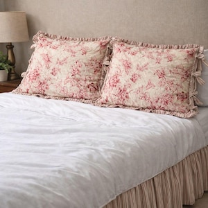 May include: Two square throw pillows with a floral pattern in shades of pink and cream rest on a white bed. The pillows have ruffled edges and are placed on a white comforter. The bed skirt is striped in pink and white.