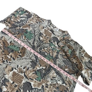 Puede incluir: Una camiseta con estampado de camuflaje con una mezcla de dise&ntilde;os de hojas y &aacute;rboles marrones, verdes y grises. Una cinta m&eacute;trica rosa est&aacute; colocada sobre la camiseta, mostrando las medidas en cent&iacute;metros. La camiseta tiene cuello redondo y mangas largas.