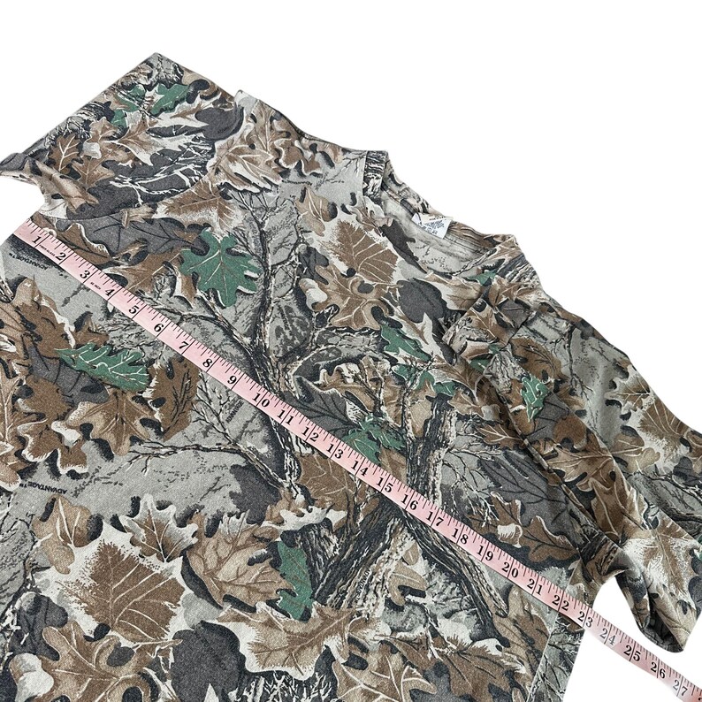 Puede incluir: Una camiseta con estampado de camuflaje con una mezcla de dise&ntilde;os de hojas y &aacute;rboles marrones, verdes y grises. Una cinta m&eacute;trica rosa est&aacute; colocada sobre la camiseta, mostrando las medidas en cent&iacute;metros. La camiseta tiene cuello redondo y mangas largas.