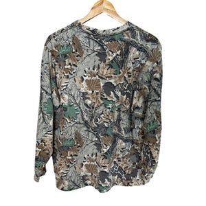 Puede incluir: Camiseta de camuflaje de manga larga con cuello redondo. La camiseta presenta un estampado de hojas y ramas en tonos verdes, marrones y grises. La camiseta est&aacute; colgada de una percha de madera sobre un fondo blanco.