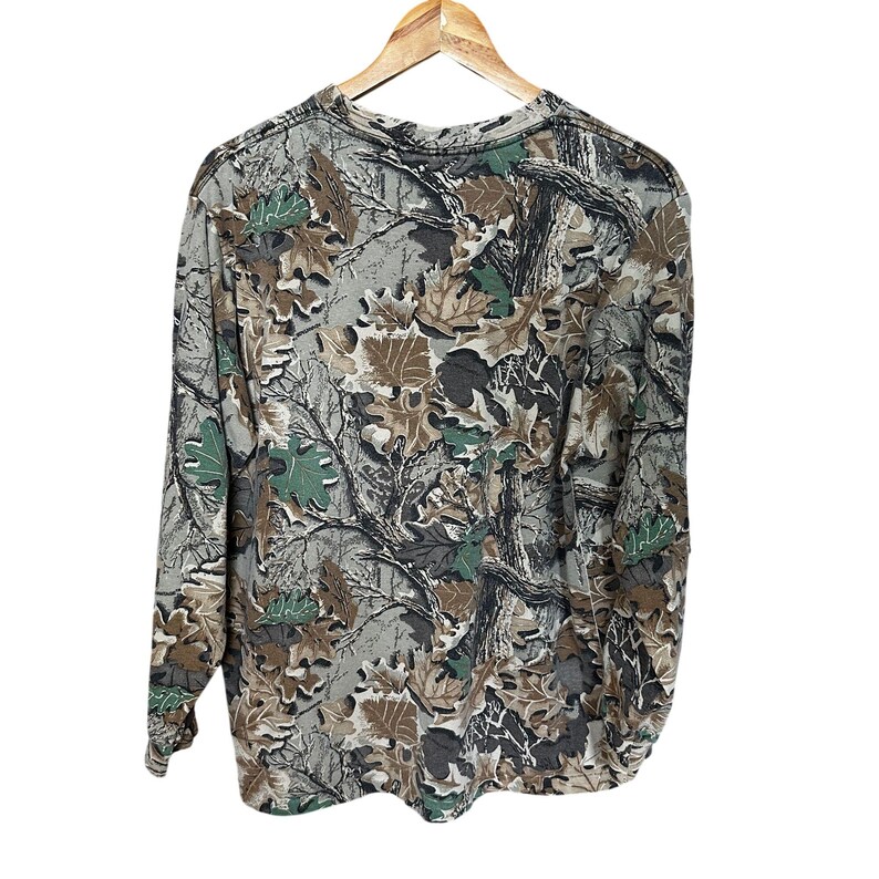 Puede incluir: Camiseta de camuflaje de manga larga con cuello redondo. La camiseta presenta un estampado de hojas y ramas en tonos verdes, marrones y grises. La camiseta est&aacute; colgada de una percha de madera sobre un fondo blanco.