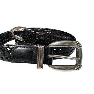May include: Black leather belt with a woven design. The belt features a silver-colored buckle with decorative scrollwork. The belt is designed with a classic style and is suitable for various outfits.