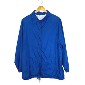 May include: A bright blue coach's jacket hanging on a wooden hanger. The jacket has a button-up front, two front pockets, and a drawstring at the bottom. The jacket is made of a lightweight material.