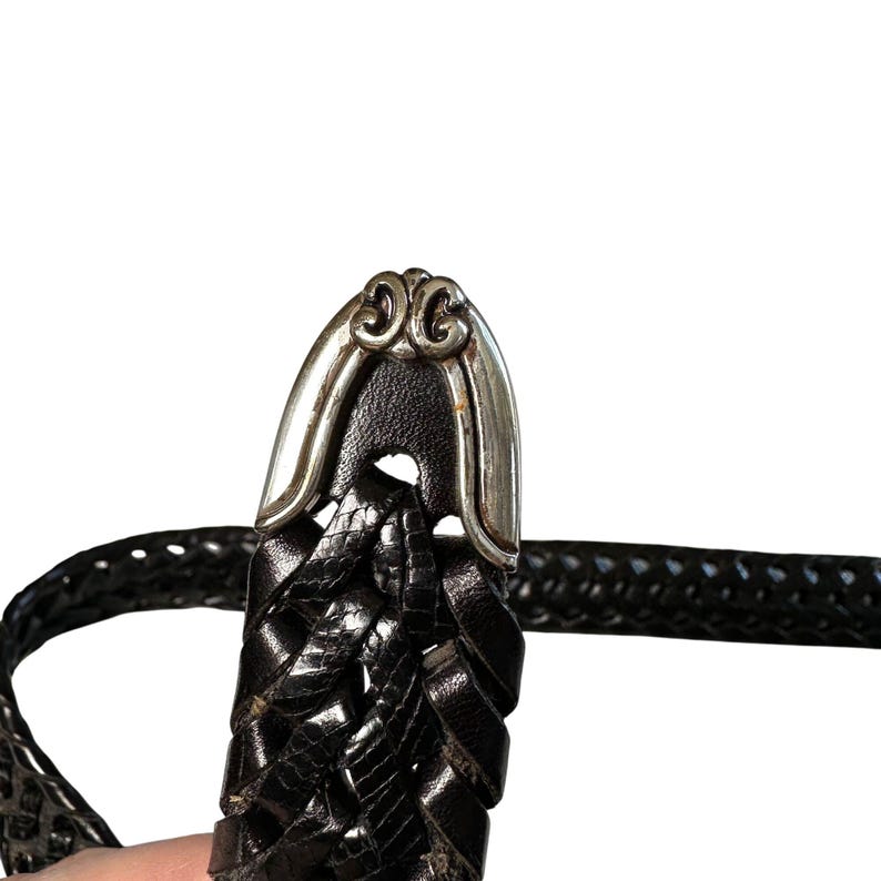 May include: Close-up of a black leather belt with a silver-toned buckle. The belt features a woven design and a decorative buckle with an ornate pattern. The belt is a classic accessory.