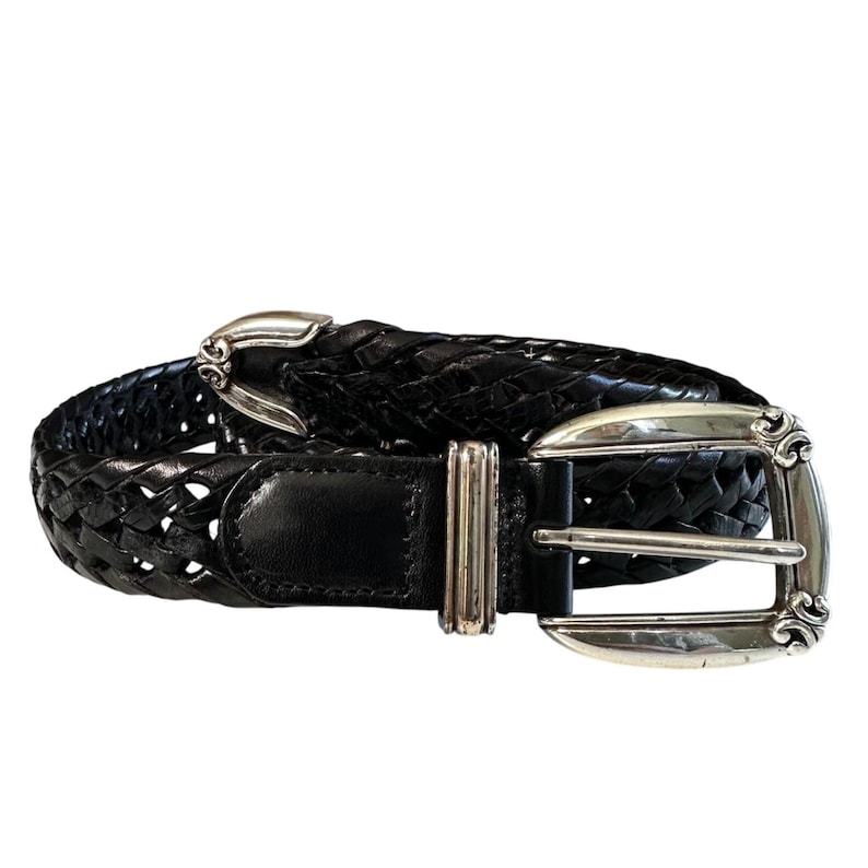 May include: Black woven leather belt with a silver-toned buckle and keeper. The belt features a braided design and a rectangular leather tab near the buckle. The buckle has an ornate design.