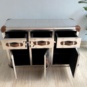 May include: A silver metal cabinet with three drawers and two doors. The cabinet has brown leather handles and corner accents. The drawers and doors are open, revealing black interiors. The cabinet is a piece of furniture.