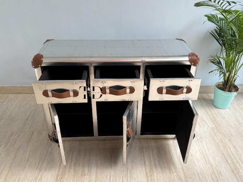 May include: A silver metal cabinet with three drawers and two doors. The cabinet has brown leather handles and corner accents. The drawers and doors are open, revealing black interiors. The cabinet is a piece of furniture.