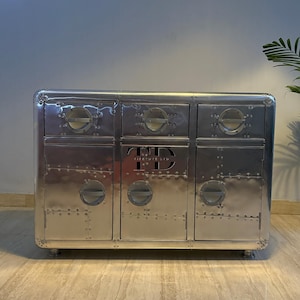 Puede incluir: Un mueble plateado con seis cajones y puertas. El mueble presenta un diseño de metal remachado con detalles circulares y el texto "TD Furniture Ltd" grabado en la parte delantera. El mueble se apoya en patas cortas y redondeadas.