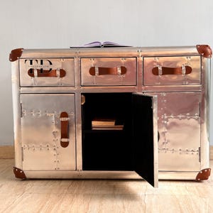 May include: A silver metal cabinet with three drawers and two doors. The cabinet has brown leather handles and corner accents. The open door reveals a black interior with books. A book rests on top of the cabinet.