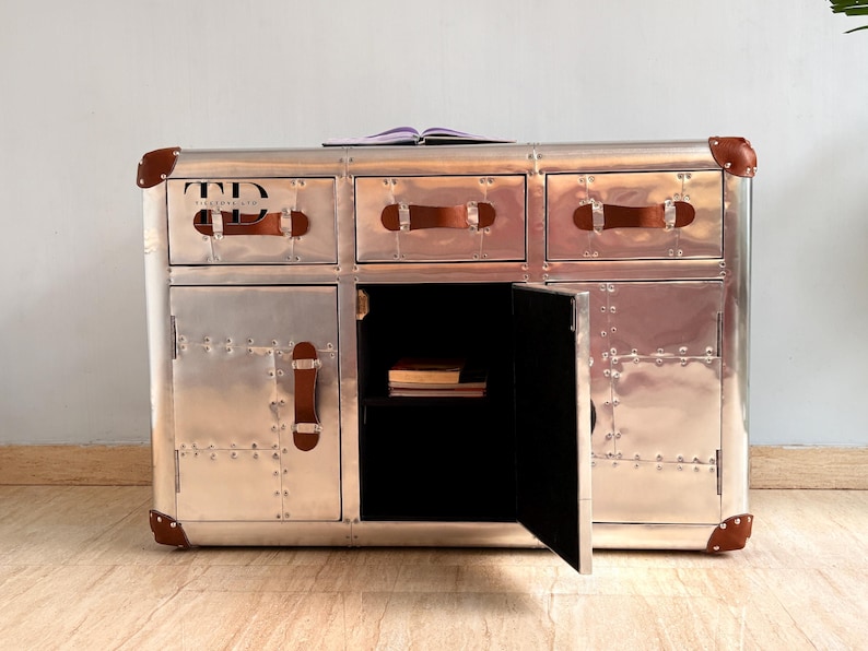 May include: A silver metal cabinet with three drawers and two doors. The cabinet has brown leather handles and corner accents. The open door reveals a black interior with books. A book rests on top of the cabinet.