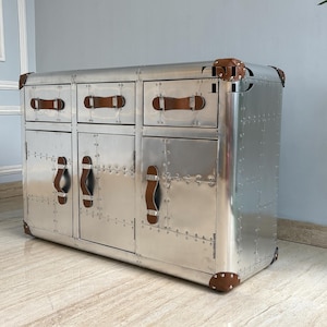 May include: A silver metal cabinet with three drawers and two doors. The cabinet has brown leather handles and corner accents. The surface is covered in rivets, giving it an industrial or vintage aesthetic. The cabinet is a storage unit.