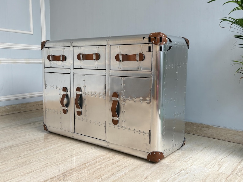 May include: A silver metal cabinet with three drawers and two doors. The cabinet has brown leather handles and corner accents. The surface is covered in rivets, giving it an industrial or vintage aesthetic. The cabinet is a storage unit.