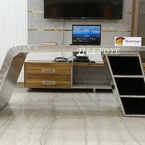 Könnte beinhalten: Ein silberner Metallschreibtisch mit einem schwarzen Regal und einem Holzkasten mit zwei Schubladen. Der Schreibtisch hat ein einzigartiges Design mit einer abgerundeten Kante und einer erhöhten Lippe. Der Schreibtisch steht an einer Wand mit einem Fernseher und einem Laptop darauf.