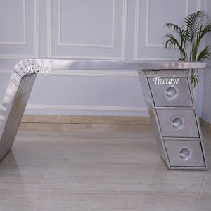 May include: A silver metal desk with a unique curved design and three drawers. The desk has a distressed finish and rivets throughout.