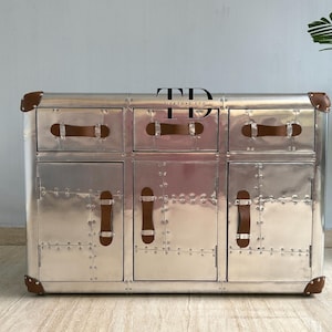 Puede incluir: Un mueble de metal plateado con seis cajones y puertas. El mueble tiene tiradores y esquinas de cuero marrón. La superficie está cubierta de remaches. Es un mueble.