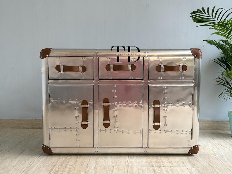 May include: A silver metal cabinet with six drawers and doors. The cabinet has brown leather handles and corner accents. The surface is covered in rivets. The cabinet is a piece of furniture.