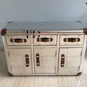 May include: Silver-colored cabinet with three drawers and two doors. The cabinet has brown leather handles and corner accents. The surface is covered in rivets. The letters "TD" are visible on the top.