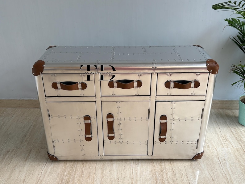May include: Silver-colored cabinet with three drawers and two doors. The cabinet has brown leather handles and corner accents. The surface is covered in rivets. The letters "TD" are visible on the top.