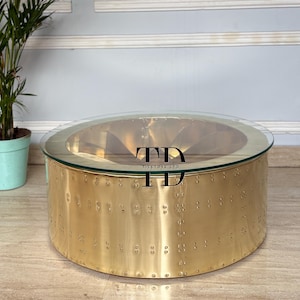 Könnte beinhalten: Runder Couchtisch mit klarer Glasplatte und Messingfarbenem Metallfuß. Der Fuß hat ein genietetes Design und eine polierte Oberfläche. Ein stilvolles Möbelstück für das Wohnzimmer.