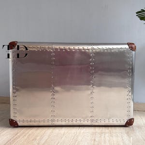 May include: A silver metal cabinet with a riveted design, featuring brown leather corner accents. The cabinet has a rectangular shape and a polished, reflective surface. A modern storage solution.