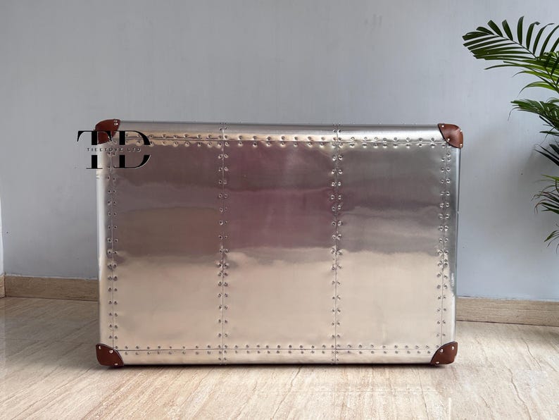 May include: A silver metal cabinet with a riveted design, featuring brown leather corner accents. The cabinet has a rectangular shape and a polished, reflective surface. A modern storage solution.