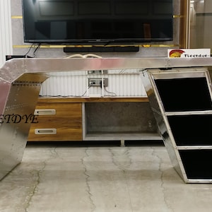 Könnte beinhalten: Ein silberner Metallschreibtisch mit einem einzigartigen Design, das ein schräges Regal mit drei Fächern aufweist. Der Schreibtisch hat eine silberne Tischplatte und ein silbernes Gestell mit einem geschwungenen Design. Der Schreibtisch steht vor einem Holzschrank mit zwei Schubladen.