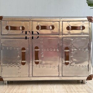 May include: A silver-colored cabinet with three drawers and two doors. The cabinet has brown leather handles and corner accents. The metal surface is covered in rivets. The cabinet sits on short, silver legs.