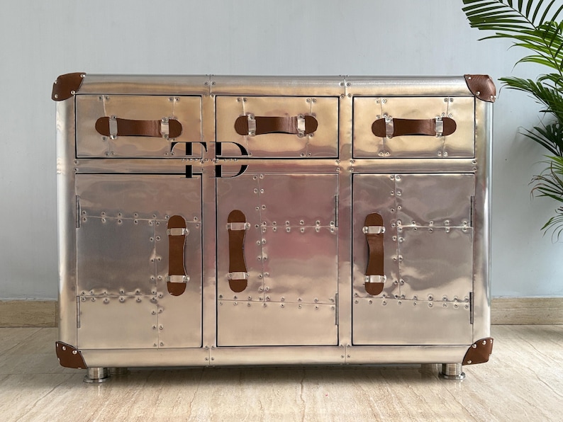 May include: A silver-colored cabinet with three drawers and two doors. The cabinet has brown leather handles and corner accents. The metal surface is covered in rivets. The cabinet sits on short, silver legs.