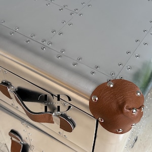 May include: A silver metal chest with a riveted design and brown leather handles and accents. The chest has a rounded brown leather knob with silver studs. The chest has the text "FIRSTDYE LTD" on the front.