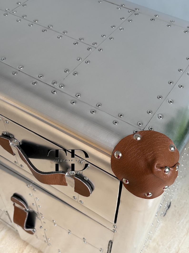 May include: A silver metal chest with a riveted design and brown leather handles and accents. The chest has a rounded brown leather knob with silver studs. The chest has the text "FIRSTDYE LTD" on the front.