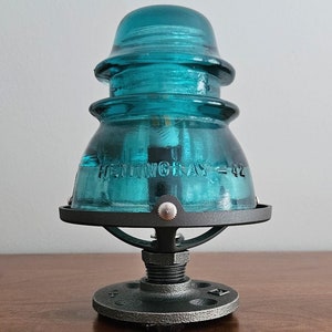 Pu&ograve; includere: Una lampada con isolatore in vetro Hemingray-42 color turchese. Il vetro &egrave; montato su una base in metallo nero con un piede rotondo in stile industriale. Il testo "HEMINGRAY = 42" &egrave; visibile sul vetro. Un oggetto decorativo unico.