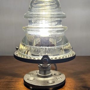 Pu&ograve; includere: Una lampada in vetro trasparente dal design industriale vintage. La lampada presenta un paralume in vetro isolante con la scritta "HEMINGRAY-42" e una base in metallo. La base &egrave; realizzata con una flangia metallica e raccordi per tubi. La lampada &egrave; accesa, emettendo un bagliore caldo.