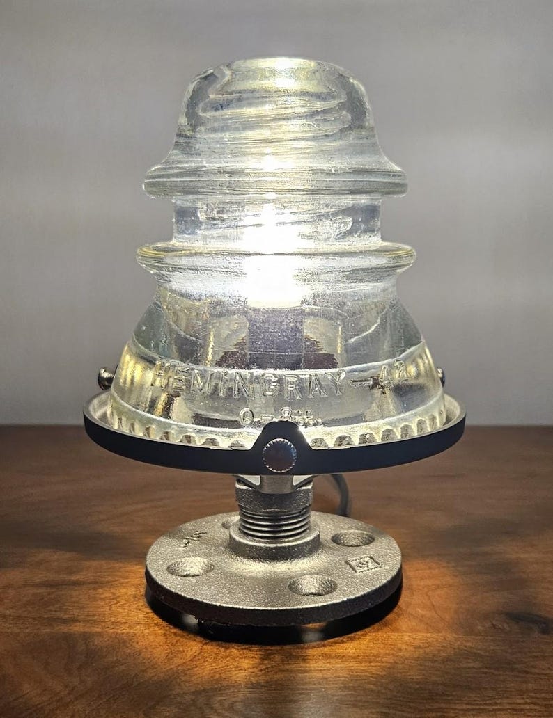 Pu&ograve; includere: Una lampada in vetro trasparente dal design industriale vintage. La lampada presenta un paralume in vetro isolante con la scritta "HEMINGRAY-42" e una base in metallo. La base &egrave; realizzata con una flangia metallica e raccordi per tubi. La lampada &egrave; accesa, emettendo un bagliore caldo.