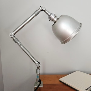 May include: A silver-colored adjustable desk lamp with a domed shade. The lamp has a clamp base and a flexible arm with multiple joints. The lamp is positioned on a wooden surface next to a notebook.