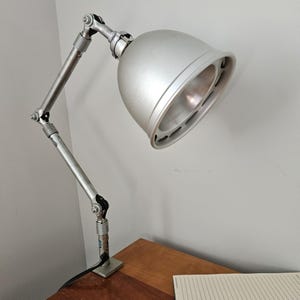 May include: Silver-toned adjustable desk lamp with a domed shade. The lamp features a flexible arm and a square base. The lamp is positioned on a wooden surface, with a notebook visible in the foreground. The lamp is plugged in.