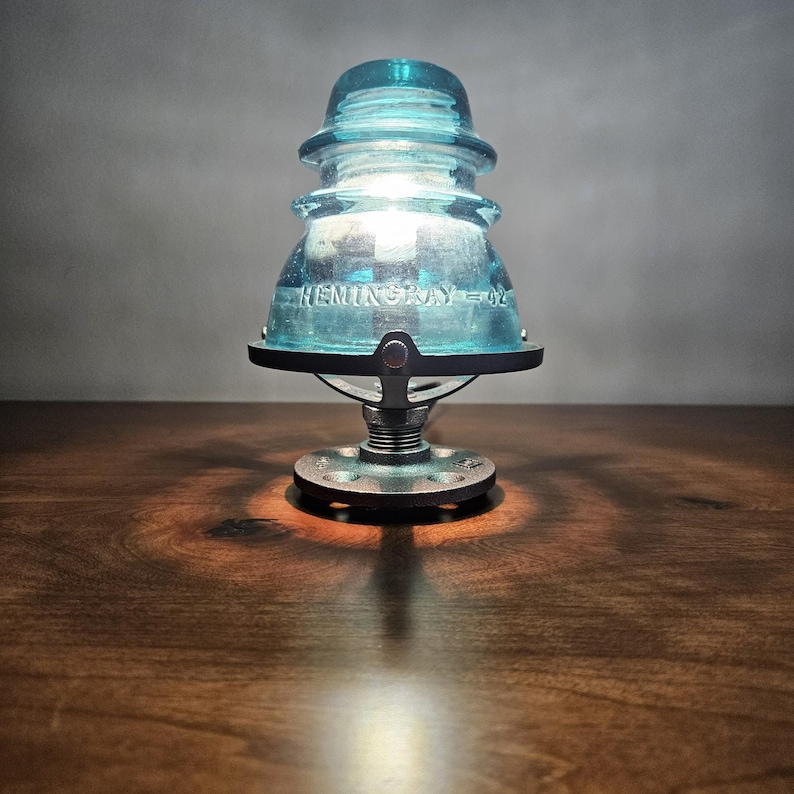 Pu&ograve; includere: Una lampada da tavolo unica realizzata con un isolatore in vetro blu vintage. La lampada ha una base in metallo e emette una luce calda. L'isolatore &egrave; contrassegnato con la scritta "HEMINGRAY = 02".