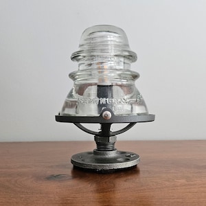 Pu&ograve; includere: Lampada con isolatore in vetro trasparente con base e hardware in metallo nero. La lampada presenta il testo "HEMINGRAY-18" sul vetro. La lampada &egrave; su una superficie di legno.