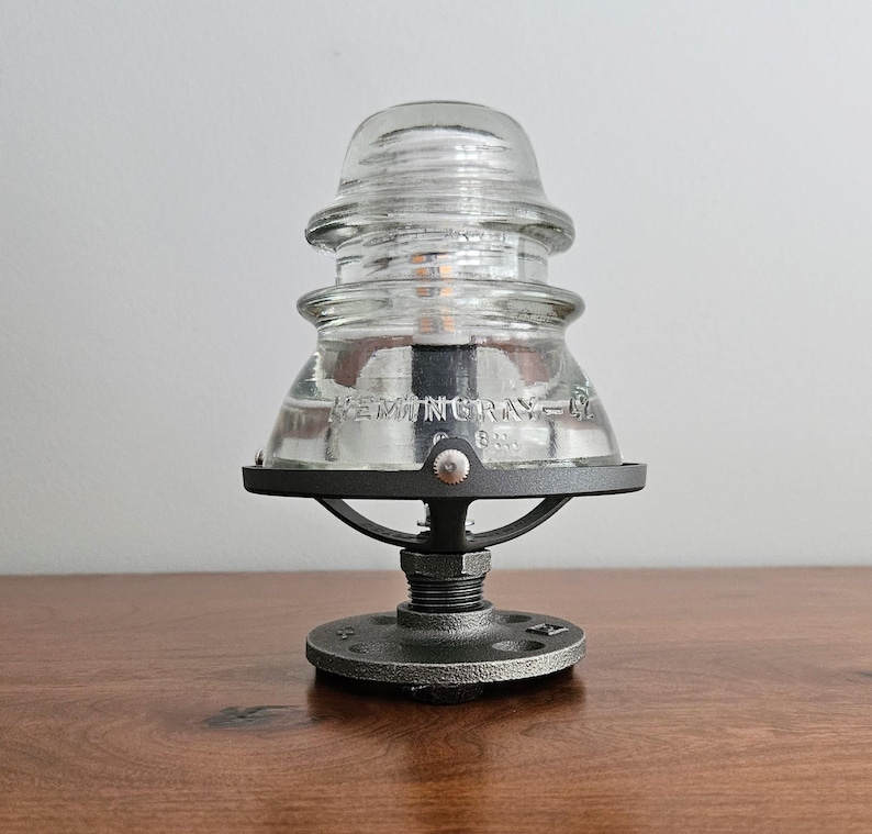 Pu&ograve; includere: Lampada con isolatore in vetro trasparente con base e hardware in metallo nero. La lampada presenta il testo "HEMINGRAY-18" sul vetro. La lampada &egrave; su una superficie di legno.